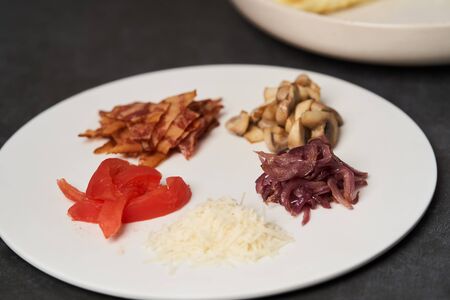 Traditional breakfast omelette with bacon, tomatoes, mushrooms, onion and cheese on a white plate on dark background with copy space, close-up. Egg omelette with parsley dill herbs.の写真素材