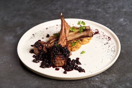 Lamb ribs with baked potatoes on a white plate, close-up. Barbecued lamb ribs with potatoes and blueberries sauce. Luxury restaurant menu foodの写真素材