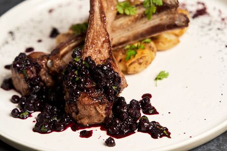 Lamb ribs with baked potatoes on a white plate, close-up. Barbecued lamb ribs with potatoes and blueberries sauce. Luxury restaurant menu foodの写真素材
