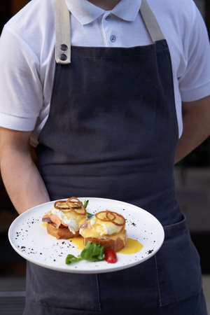Waiter holding breakfast tray with Eggs Benedict with smoked salmon, poached eggs. Restaurant breakfast menu with toast, avocado, poached eggs. Hotel serviceの写真素材
