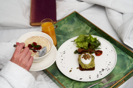 Woman reading book while having breakfast in bed. Woman in bathrobe having breakfast in hotel bed,...