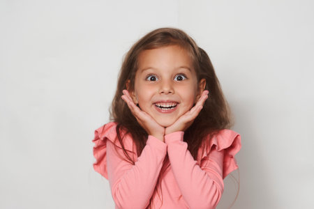 Portrait of surprised little girl against white background with copy space. Beautiful girl holds cheeks by hand. Expressive facial expressionsの写真素材