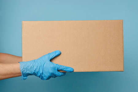Delivery man holding cardboard box on blue background with copy space, close-up. Online shopping and...