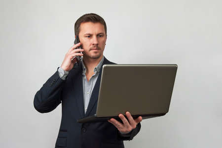 Young businessman using laptop and phone on white background with copy space. Portrait of successful man holding laptop computer and smartphone in hands, close-upの写真素材