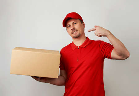 Young delivery man in red uniform standing with parcel box on white background. Portrait of handsome...