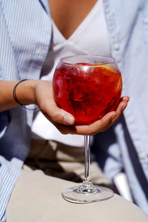 Woman hand holding glass with red cocktail, close-up. Woman drink fresh cocktail with ice, outdoorsの写真素材