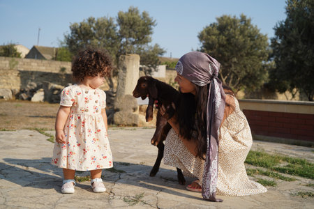 Happy mother and daughter playing with sheep outdoor. Young woman with baby girl and little lamb