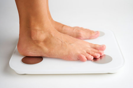 Woman legs stepping on floor scales, close-up. Female bare feet standing on scales, white background. Diet and overweight concept.の写真素材