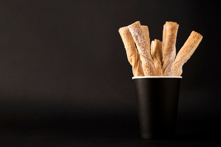 Paper coffee cup and Puff pastry sticks with cinnamon on black background with copy space. Take away coffee and breadsticks sprinkled with sugar, close-upの写真素材