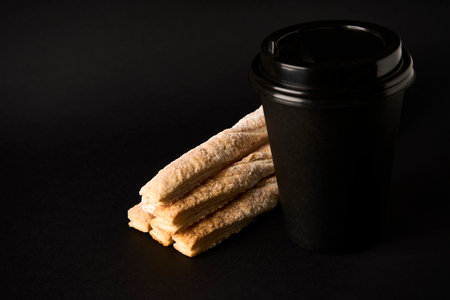 Paper coffee cup and Puff pastry sticks with cinnamon on black background with copy space. Take away coffee and breadsticks sprinkled with sugar, close-upの写真素材
