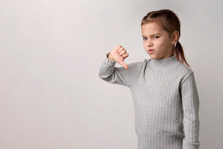 Baby girl giving thumbs down hand gesture, white background with copy space, close-up. Portrait unhappy child girl with long brown hair, wearing gray dress, showing dislike gestureの写真素材