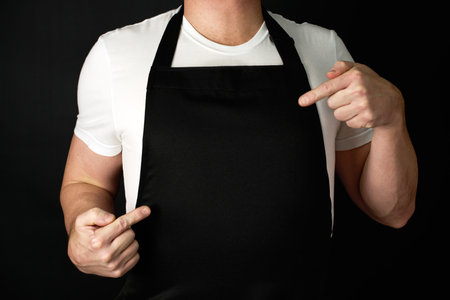 Chef pointing on black apron, close-up. Male chef wearing apron, standing on a black background. Can be used for restaurant menuの写真素材