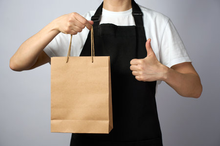 Woman holding take away paper bag and showing thumbs up gesture over white background, close-up. Takeaway food and delivery serviceの写真素材