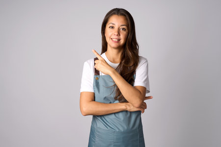 Woman with apron pointing to the side to present a product over isolated grey background with copy spaceの写真素材