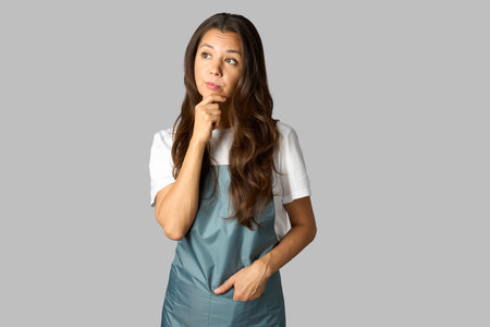Portrait of thinking woman barista standing with hand on chin. Woman wearing apron and white shirt, white backgroundの写真素材