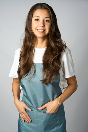 Friendly female barista in a apron standing with hands in pockets, smiling against a light gray backgroundの写真素材