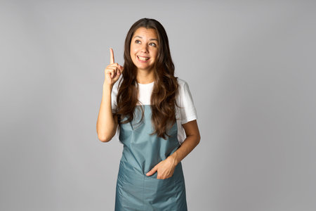 Happy smiling woman wearing apron, standing with finger pointing up. Female having an ideaの写真素材