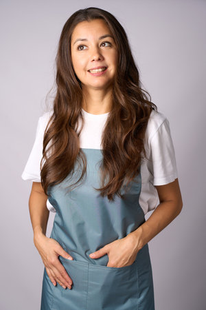 Friendly female barista in a apron standing with hands in pockets, smiling against a light gray backgroundの写真素材