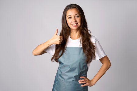 Happy young woman in apron showing thumb up on grey background with copy spaceの写真素材