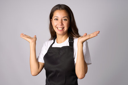 Friendly waitress, female barista in black apron introduce new product. Young woman wearing black apron and white shirt standing with open hands on a white background with copy spaceの写真素材