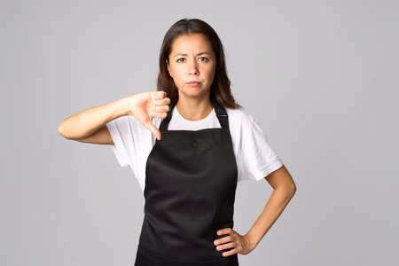 Young woman wearing black apron with angry face, negative sign showing dislike with thumbs downの写真素材