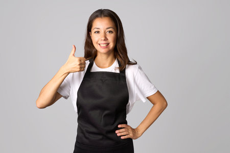 Friendly woman in apron showing thumb up on grey background with copy spaceの写真素材