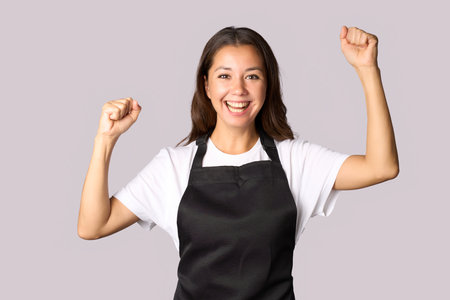 Portrait of cheerful woman wear apron with raised hands, celebrating successの写真素材