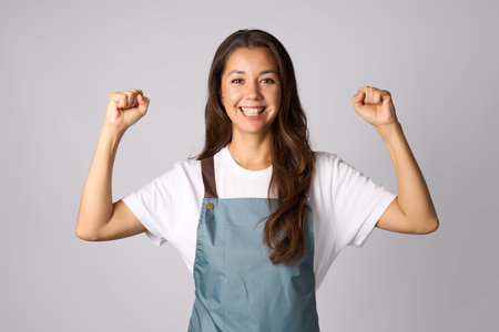 Portrait of cheerful woman wear apron with raised hands, celebrating successの写真素材