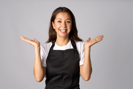 Friendly waitress, female barista in black apron introduce new product. Young woman wearing black apron and white shirt standing with open hands on a white background with copy spaceの写真素材