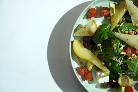 A top view of a salad in a light blue bowl. The salad contains greens, tomatoes, pears, and pine...