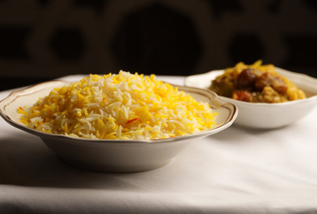 Fluffy basmati rice infused with saffron, displayed in a decorative white bowl. A stew or curry is visible in a slightly blurred bowl in the backgroundの写真素材