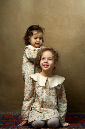 Portrait of Two Sisters in Matching Outfits. Two adorable sisters in vintage-style floral dresses pose near each other, one sitting and one standing behindの写真素材