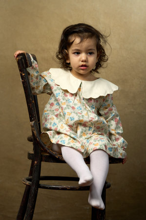 Toddler Girl Sitting on Antique Chair. A cute girl in a floral dress and tights sits on a rustic...