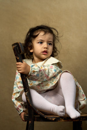 Toddler Girl Sitting on Antique Chair. A cute girl in a floral dress and tights sits on a rustic wooden chair, looking back over her shoulder with a curious expressionの写真素材