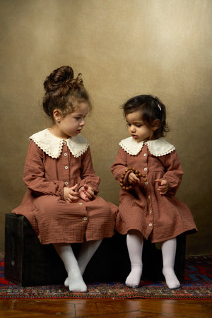 Two Sisters Looking at Each Other. Two young sisters in matching brown dresses with lace collars sit on separate black blocks, looking at each other with gentle expressionsの写真素材