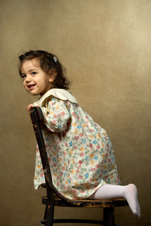 Joyful Little Girl on Wooden Chair. A cute girl in a vintage-style floral dress stands on an old wooden chair, looking and laughing with delightの写真素材