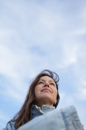 Low-Angle Portrait of a Woman Looking Up at the Sky. A woman with long brown hair and freckles, wearing a light blue denim jacket, is seen from a low angle, looking upwards towards a bright skyの写真素材