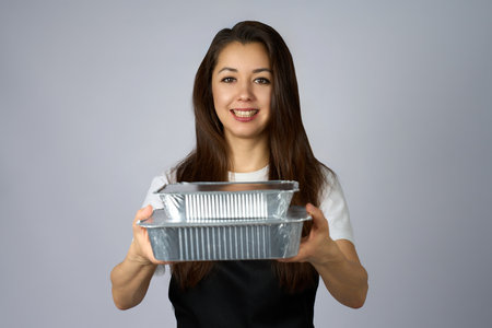 Portrait of a cheerful woman wearing an apron and holding aluminum food containers. Concept of food delivery service, meal prep, catering business, or home-cooked meals to-goの写真素材