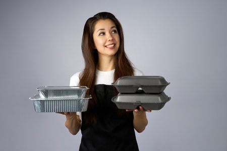 Young Woman Presenting Takeaway Containers. A smiling woman holds a stack of silver foil and black polystyrene container in the other, presenting them outwards against a plain grey backdropの写真素材