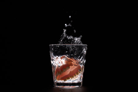 Dentures falling into glass of water with splash, black background with copy space. Cleanliness, dental hygiene, and night care routines for removable false teeth. Concept oral care, elderly hygieneの写真素材