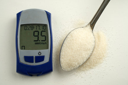 A close-up, high-angle shot of a digital glucose meter displaying a blood sugar reading next to a spoonful of white granulated sugar on a light blue surfaceの写真素材