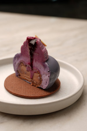 A modern purple mousse pastry on a chocolate biscuit base, topped with piped cream and almond...