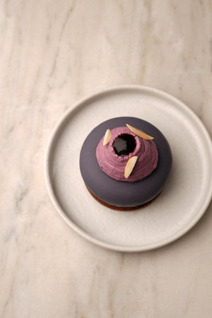 A modern purple mousse pastry on a chocolate biscuit base, topped with piped cream and almond...