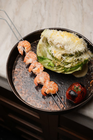 Skewer of grilled shrimp served alongside a wedge of Caesar salad topped with shredded cheese and a roasted tomato on a dark, textured plateの写真素材