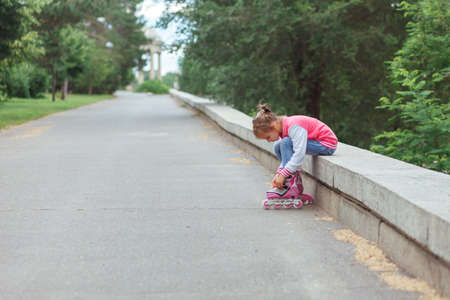 Little girl wearing jeans and jacket sitting on parapet and putting on rollers in the park in summer day.の写真素材
