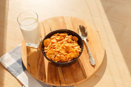 Sweet cornflakes breakfast cereal in the bowl with glass of milk on the wood table and spoon. Scene lit with direct sun rays. Horizontal image. Selective focusの写真素材