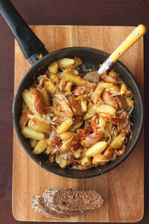 Fried potatoes in a frying pan with a fork in it and with two pieces of dark rye bread on the cutting boardの写真素材