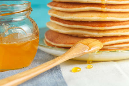 Close up of stack of pancakes with pouring honey on plate with wooden spoon and jar with honey. Two drops of honey drip on napkin. Concept of shrovetide treats, close-up view. Selective focusの写真素材