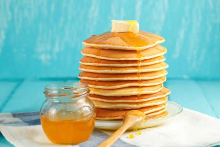 Stack of pancakes with pouring honey and piece of butter on plate with wooden spoon and jar with honey. Two drops of honey drip on napkin. Concept of shrovetide treats, close-up view. Selective focusの写真素材