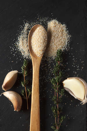 Vertical photo of garlic powder sprinkled into wooden spoon, two sprigs of thyme and thre cloves on black slate board. Selective focusの写真素材